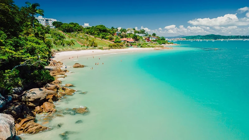 Pequena praia de águas calmas e verde-turquesa entre pedras e mata, com algumas pessoas na água e casas na encosta