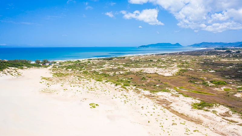 Dunas de areia branca com vegetação rasteira, o mar azul e morros no horizonte em um dia de céu azul