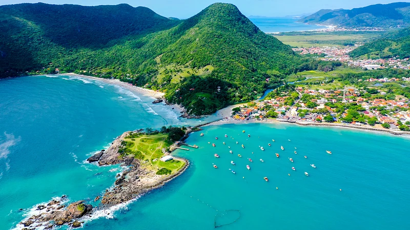 Vista aérea de uma enseada com barcos de pesca ancorados, uma vila à beira da praia e morros cobertos de mata