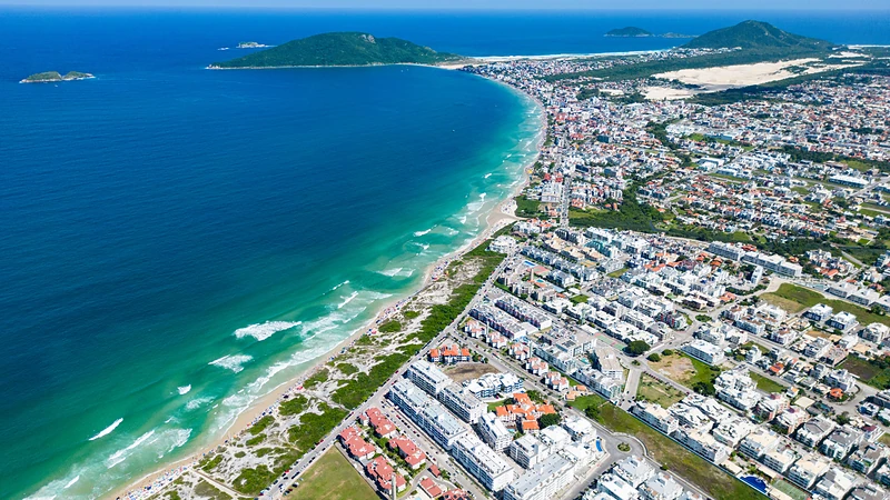 Vista aérea de uma longa praia de mar azul-turquesa e areia clara, com bairro à beira-mar, morros verdes e pequenas ilhas ao fundo